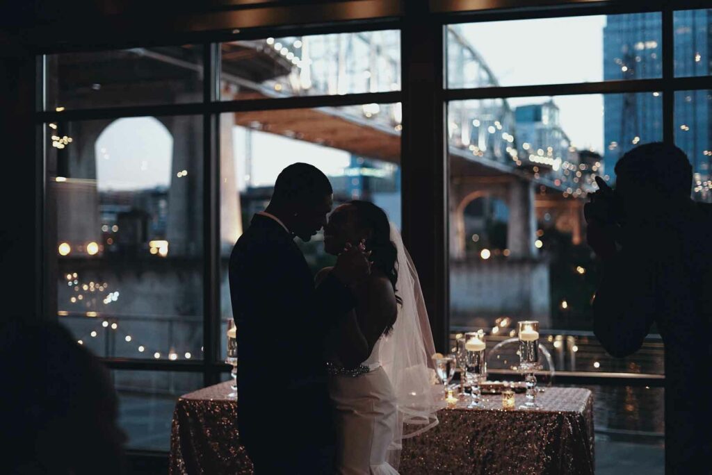 A couple is dancing at their New Year's Eve wedding indoors, with large floor-to-ceiling windows providing a view of the city. A photographer is taking their picture in the shadows.